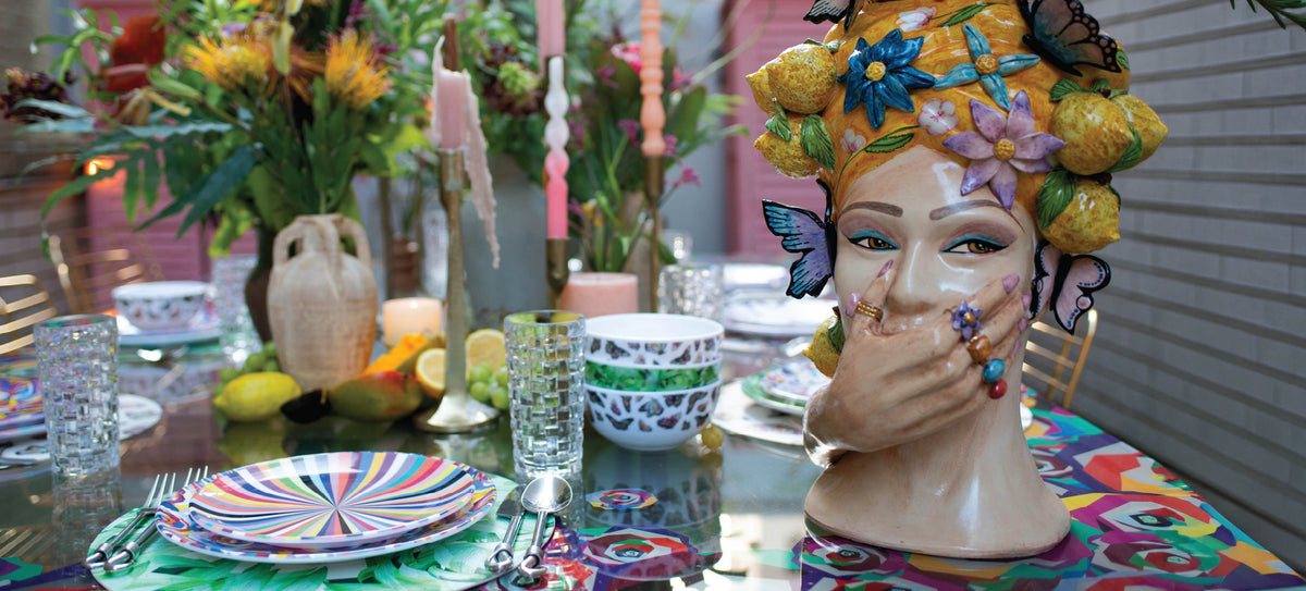Hands placing an Elizabeth Sutton Collection gift box on the grass beside styled melamine dinnerware
