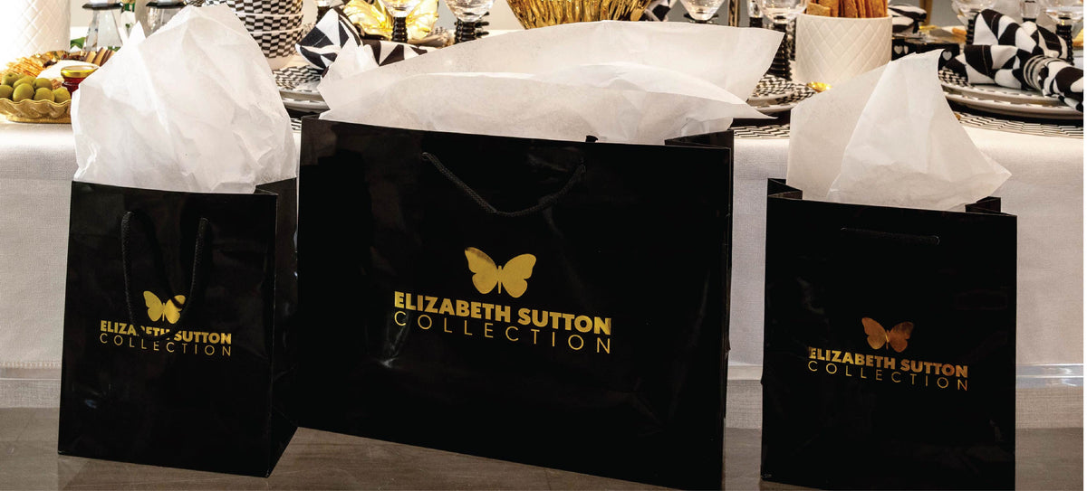 Hands placing an Elizabeth Sutton Collection gift box on the grass beside styled melamine dinnerware